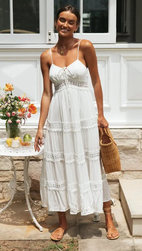 Vrouw in witte zomerjurk met kant, draagt een rieten tas en sandalen, staat voor een tafel met bloemen en fruit. Zomermode, boho-stijl.