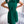 Vrouw in elegante groene jurk met plissérok en strikdetail, staand in moderne woonkamer. Mode, dameskleding, stijlvolle outfit.
