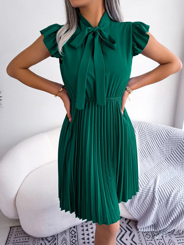 Vrouw in elegante groene jurk met plissérok en strikdetail, staand in moderne woonkamer. Mode, dameskleding, stijlvolle outfit.
