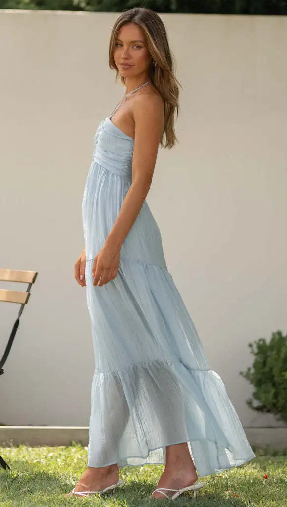 Vrouw in lichtblauwe maxi-jurk, staand naast houten stoel in tuin. Zomerse mode, elegante kleding, casual stijl, buitenomgeving.