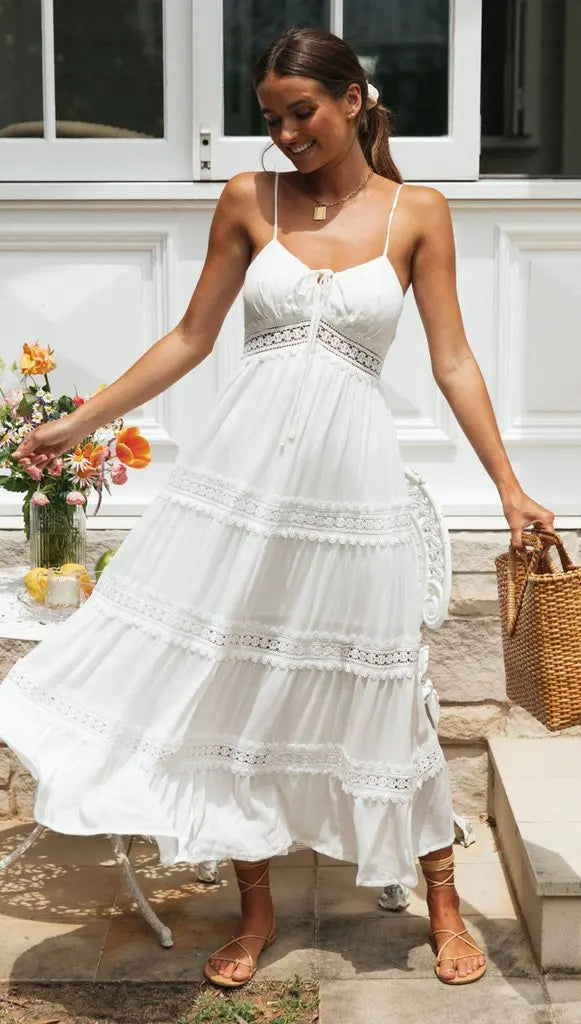 Vrouw in witte zomerjurk met kantdetails, draagt een rieten tas en sandalen, poseert buiten voor een huis met bloemen op de achtergrond.