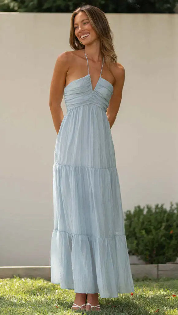 Vrouw in lichtblauwe zomerjurk met halternek, staand in tuin. Elegante maxi-jurk, perfect voor zomerse gelegenheden. Modetrends 2023.