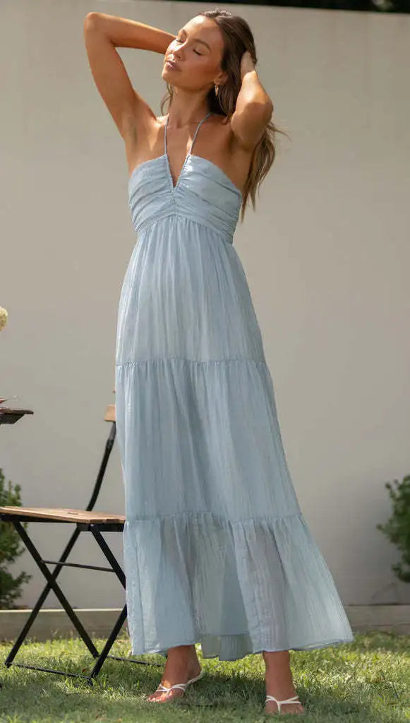 Vrouw in lichtblauwe maxi-jurk met halternek, ontspannen in tuin. Zomerse mode, elegante dameskleding, casual chique stijl.