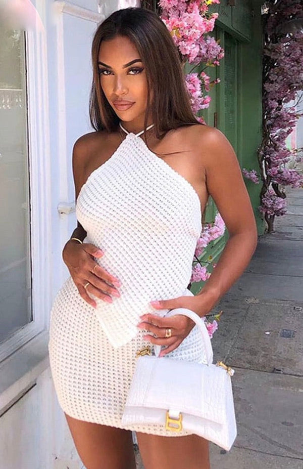 Vrouw in witte gehaakte jurk met asymmetrische halslijn, poseert buiten naast roze bloemen, houdt een witte handtas vast. Zomermode, elegante stijl.