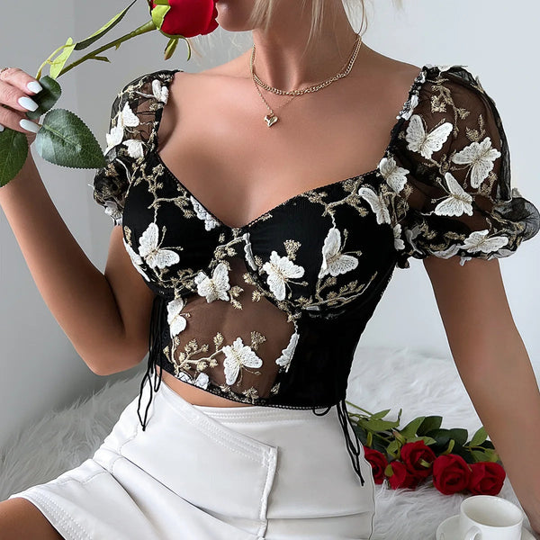 Vrouw in zwarte bloemen crop top met transparante mouwen, witte rok, houdt rode roos vast. Trendy damesmode, romantische outfit, elegante stijl.