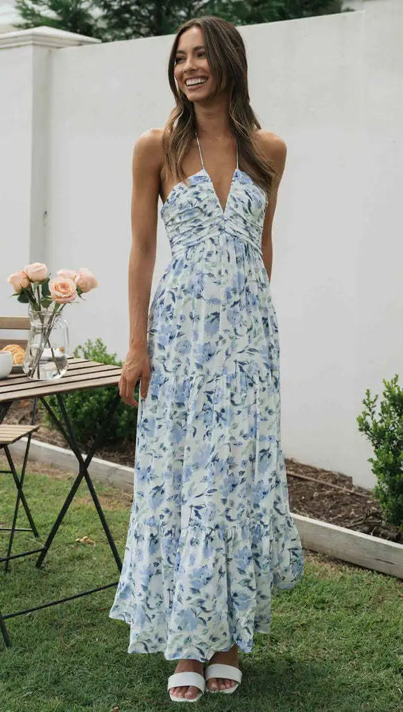 Vrouw in blauwe bloemenjurk met halternek, staand in tuin naast houten tafel met rozen. Zomermode, elegante dameskleding, casual stijl.