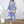 Vrouw in blauwe en witte maxi-jurk met geometrisch patroon, draagt een rieten tas en sandalen, staand op houten vloer naast witte decoratieve plant.