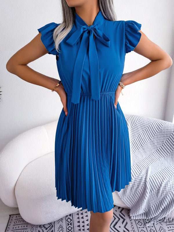 Vrouw in elegante blauwe plisséjurk met strik, staand voor witte bank. Mode, dameskleding, zomerjurk, stijlvolle outfit, trendy design.