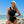 Vrouw in zwarte asymmetrische badpak met bloemen, poseert op zonnig strand. Zonnebril, blauwe lucht en oceaan op achtergrond. Zomermode, strandkleding.