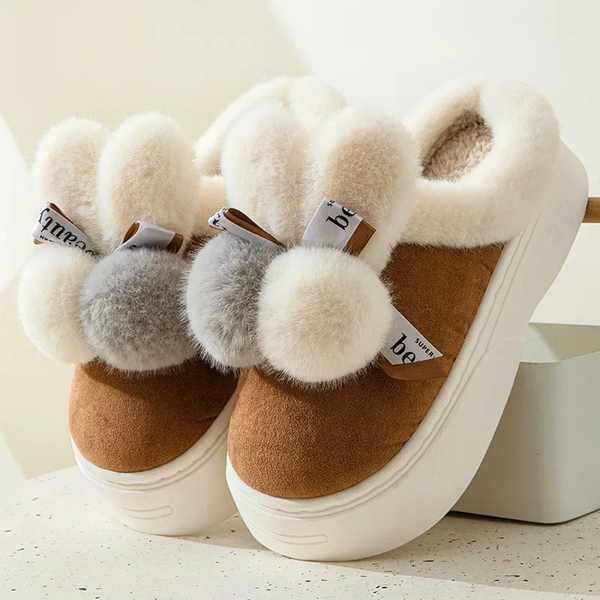 Bruine pluche pantoffels met witte en grijze pompons, dikke witte zool en zachte voering. Perfect voor wintercomfort en warme voeten.
