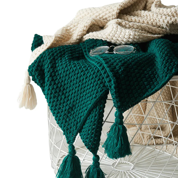 Groene gebreide deken met kwastjes op witte draadmand, naast beige deken en bril. Stijlvolle woondecoratie, gezellige textielaccessoires.