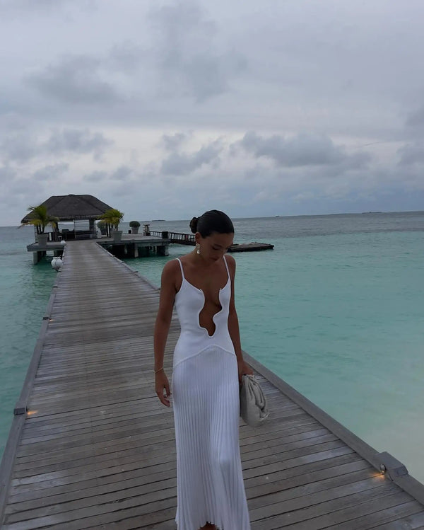 Vrouw in elegante witte jurk loopt over houten pier naar tropisch strandhuis, turquoise zee op achtergrond, bewolkte lucht. Perfect voor zomer- en strandmode.