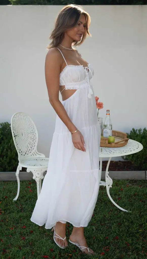 Vrouw in elegante witte zomerjurk met spaghetti bandjes, staand naast een witte tuintafel met bloemen en drankjes, op een groene grasmat.
