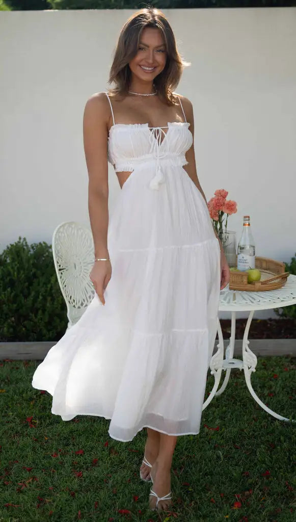 Vrouw in witte zomerjurk met spaghettibandjes, staand in tuin naast ronde tafel met bloemen en drankjes. Perfect voor zomermode en tuinfeesten.