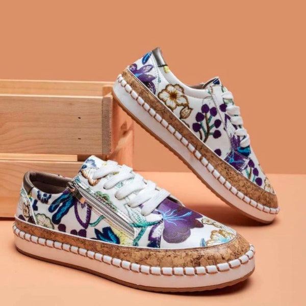 Floral damessneakers met kurkdetails en witte zolen, geplaatst op een houten kist tegen een perzikkleurige achtergrond. Trendy en duurzaam schoeisel.