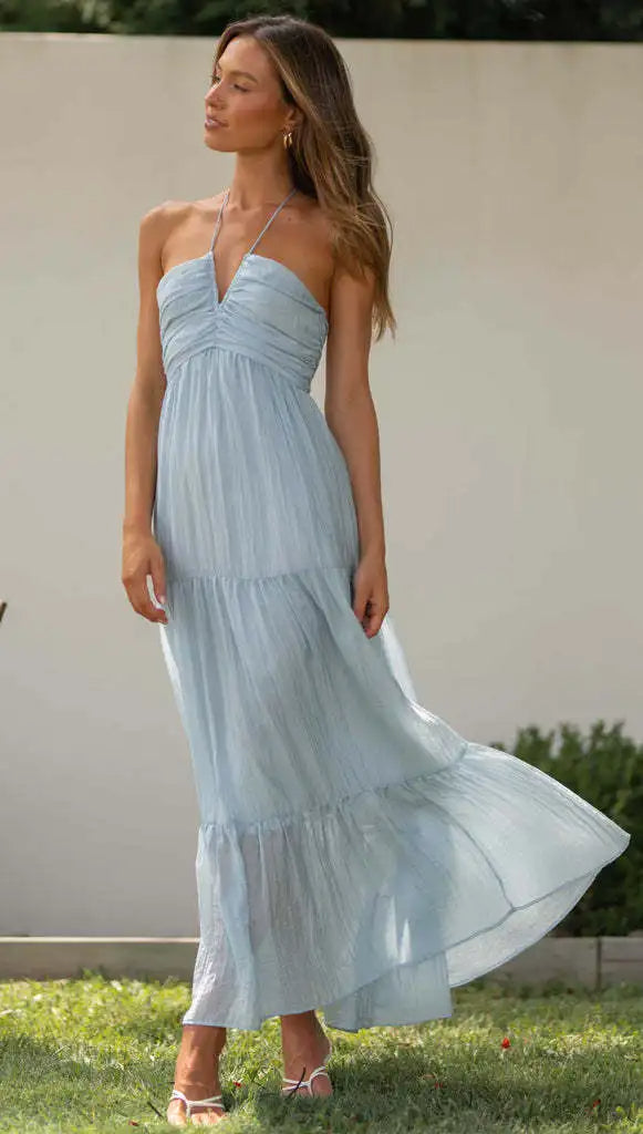 Vrouw in lichtblauwe zomerjurk met spaghettibandjes, staand in een tuin. Elegante damesmode, casual zomeroutfit, luchtige maxi-jurk.