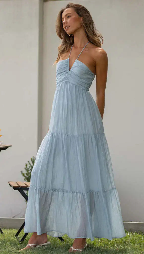 Vrouw in lichtblauwe zomerjurk met halternek, staand in tuin. Elegante maxi-jurk, perfect voor zomerse evenementen en casual stijl.