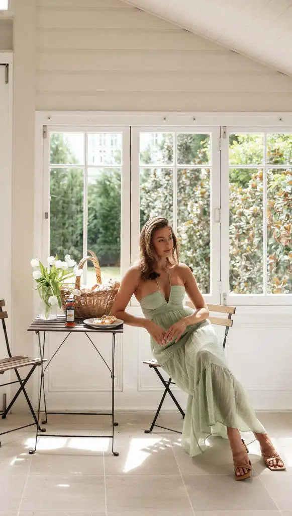 Vrouw in groene jurk zit in lichte serre naast tafel met bloemen en picknickmand; zomerse sfeer, ontspannen interieur, natuurlijke verlichting.