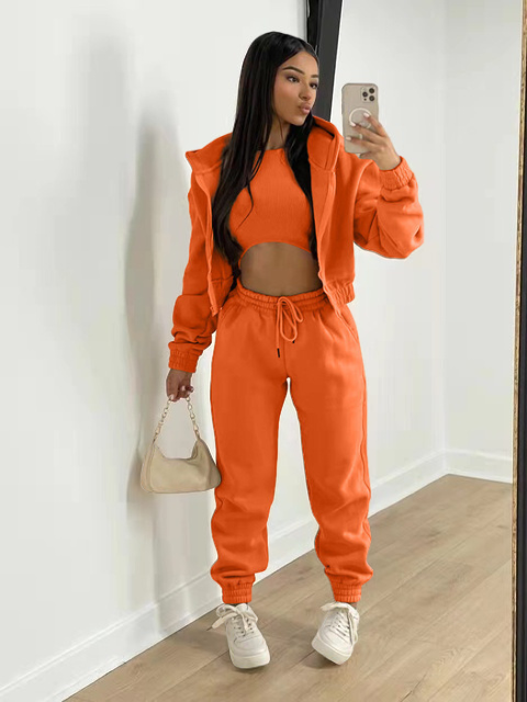 Vrouw in oranje trainingspak met crop top, houdt een beige handtas vast en maakt een selfie. Modieuze sportkleding, casual stijl, trendy outfit.