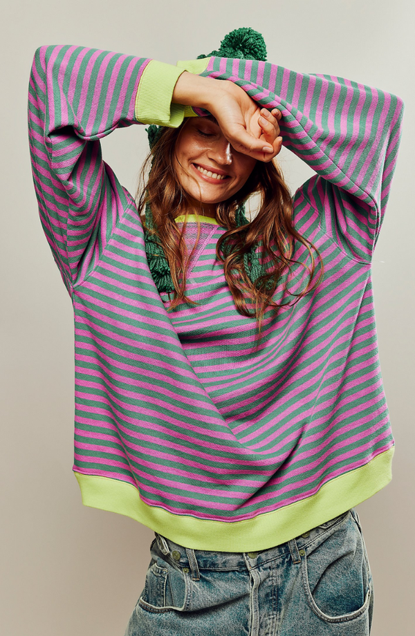 Vrouw in gestreepte trui met roze en paarse strepen, groene boorden, draagt een groene muts, lacht en poseert met armen omhoog. Casual mode.