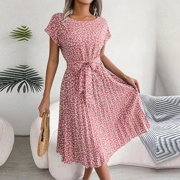 Vrouw in roze bloemenjurk met strikceintuur, houdt rieten tas vast. Zomerse outfit, casual stijl, woonkamer achtergrond.