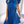Vrouw in blauwe geplooide jurk met strik en pofmouwen, draagt witte schoudertas met gouden ketting. Stijlvolle mode, elegante dameskleding.