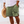 Man in witte blouse en groene korte broek met zonontwerp, strandachtergrond, strohoed in hand. Zomermode, strandkleding, casual herenkleding.
