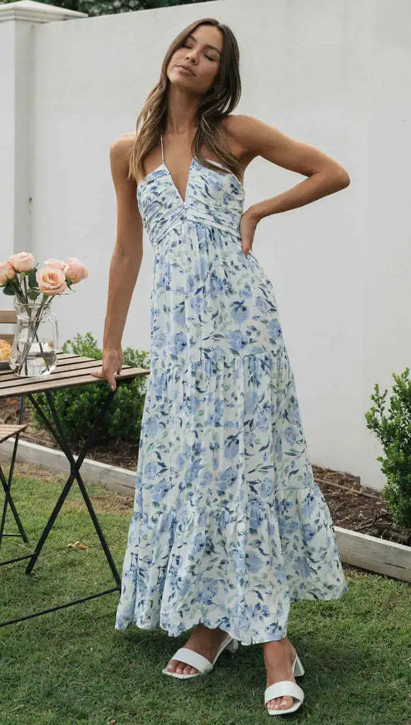 Vrouw in lange, blauwe bloemenjurk poseert buiten naast tafel met rozen; zomermode, elegante maxi-jurk, tuinfeest outfit.