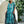 Vrouw in turquoise en zwarte gestreepte kaftan jurk, staand voor witte muur met groene planten. Zomermode, strandkleding, bohemian stijl.