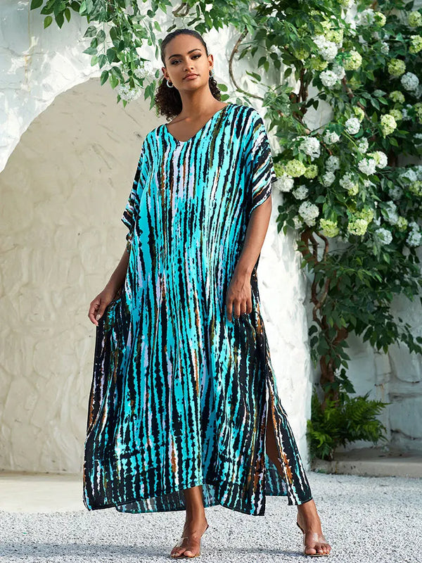Vrouw in turquoise en zwarte gestreepte kaftan jurk, staand voor witte muur met groene planten. Zomermode, strandkleding, bohemian stijl.