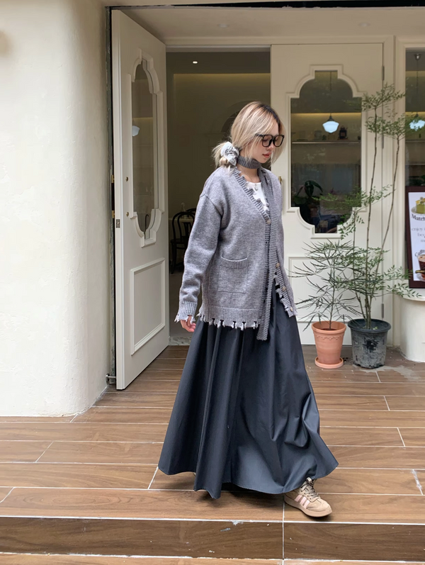 Vrouw in grijze cardigan en lange rok loopt voor winkel, modieuze herfstoutfit, casual stijl, houten vloer, plantenpot op achtergrond.