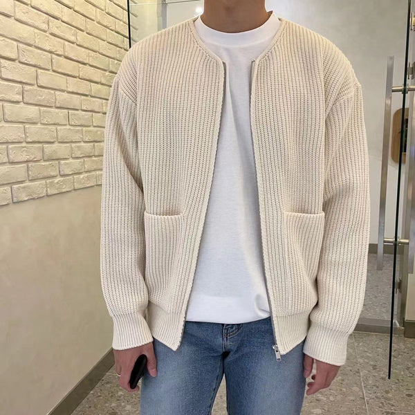 Man in crèmekleurig gebreid vest met ritssluiting, witte T-shirt en blauwe jeans, staand voor een bakstenen muur. Casual herenkleding.
