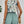 Vrouw in zomerse outfit met bloemen top en groene shorts, draagt een rieten tas. Perfecte look voor strandvakantie of casual zomeruitjes.