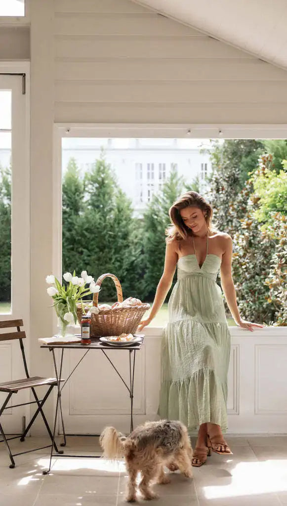 Vrouw in groene zomerjurk leunt tegen tafel met mand en tulpen, kleine hond op voorgrond. Lichte serre met tuinzicht. Zomerse sfeer.