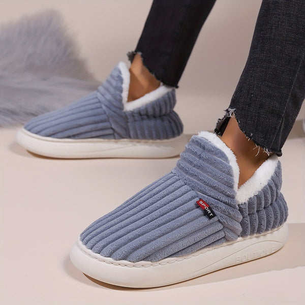 Grijze geribbelde pantoffels met witte voering, gedragen door persoon in zwarte jeans. Comfortabele winterse huisschoenen voor dames.