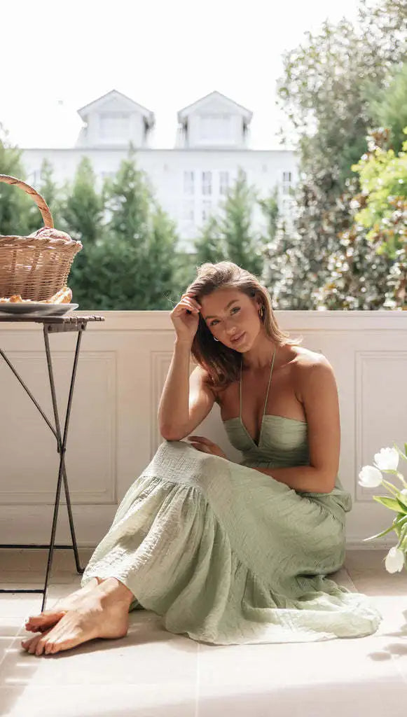 Vrouw in groene zomerjurk zittend op vloer bij raam, met mand op tafel. Lente mode, casual stijl, ontspannen sfeer, zonlicht, bloemen.