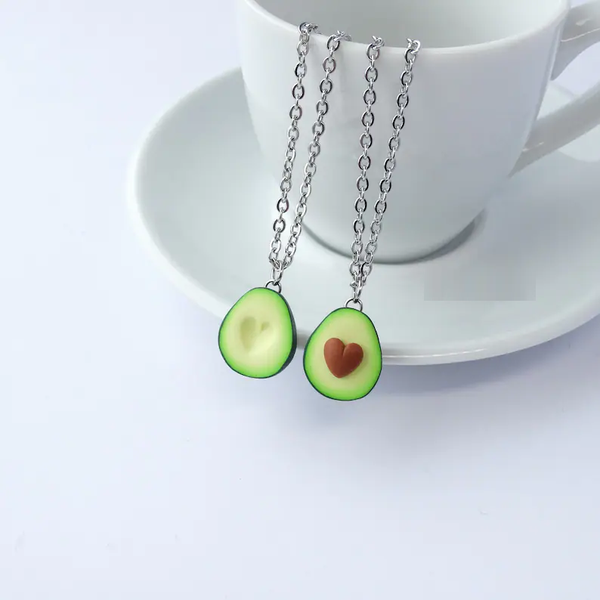 Twee zilveren kettingen met avocado-hangers, elk met een hartvormig detail, op een witte kop en schotel. Perfecte sieraden voor avocado-liefhebbers.