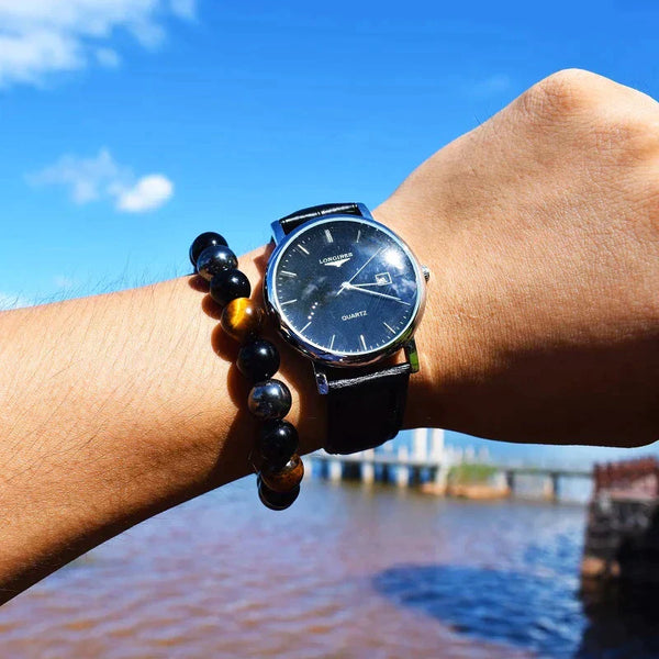 Polshorloge met zwarte wijzerplaat en leren band, gedragen naast een kralenarmband, tegen een blauwe lucht en water op de achtergrond.