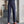 Man in blauwe blouse en grijze hoge taille broek met bruine loafers, staand naast een betonnen muur en plant. Stijlvolle herenmode, zakelijke outfit.