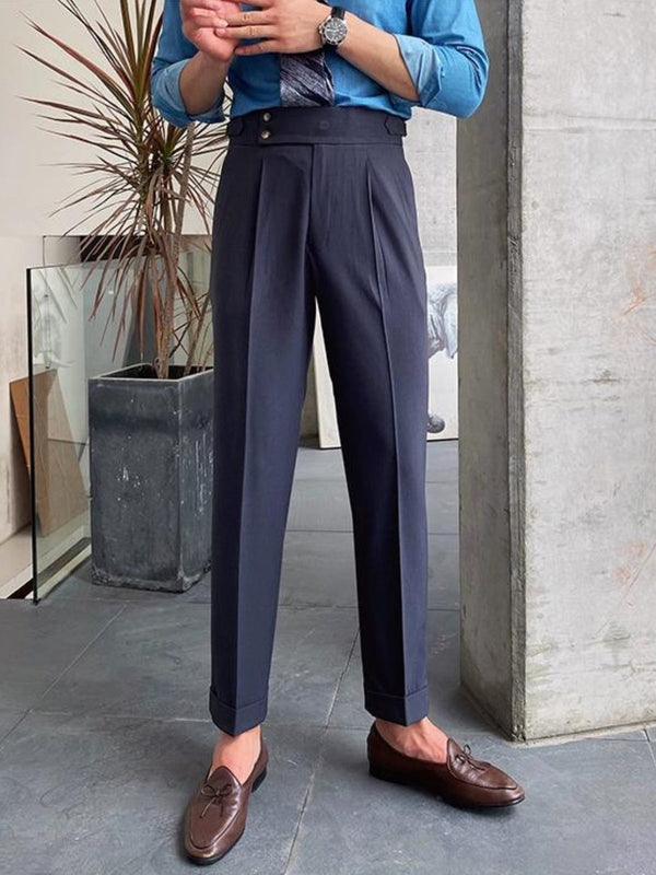 Man in blauwe blouse en grijze hoge taille broek met bruine loafers, staand naast een betonnen muur en plant. Stijlvolle herenmode, zakelijke outfit.