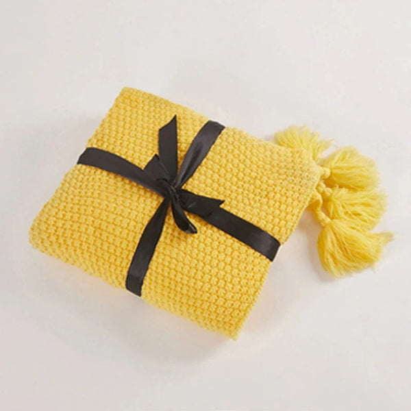 Gele gebreide deken met zwarte strik en kwastjes, perfect voor interieurdecoratie en comfort. Ideaal als cadeau of woonaccessoire.