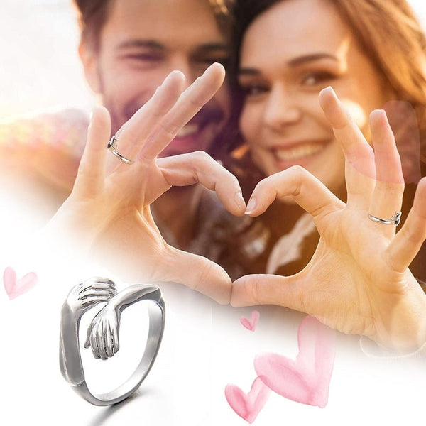 Twee mensen vormen een hart met hun handen, met op de voorgrond een zilveren ring in de vorm van een omhelzing. Romantisch cadeau, liefde, sieraden.