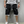 Man in witte sneakers en zwarte cargoshorts met witte touwtjes, staand tegen een grijze muur. Casual herenkleding, streetwear mode.