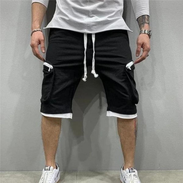 Man in witte sneakers en zwarte cargoshorts met witte touwtjes, staand tegen een grijze muur. Casual herenkleding, streetwear mode.