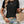 Vrouw in zwarte top en denim shorts met gevlochten rieten tas, sieraden en golvend haar tegen witte muur. Zomermode, casual outfit.