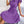 Vrouw in paarse geplooide jurk met strik, zittend op bank met geometrisch patroon deken. Modieuze dameskleding, zomerjurk, elegant design.