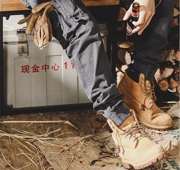 Man met bruine werkschoenen en grijze broek, zittend naast een stapel houtblokken, houdt een flesje bier vast. Outdoor kleding, casual stijl.
