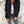 Vrouw in zwarte corduroy jas met capuchon, witte top en grijze jeans tegen betonnen muur. Modieuze herfstkleding, casual damesoutfit.