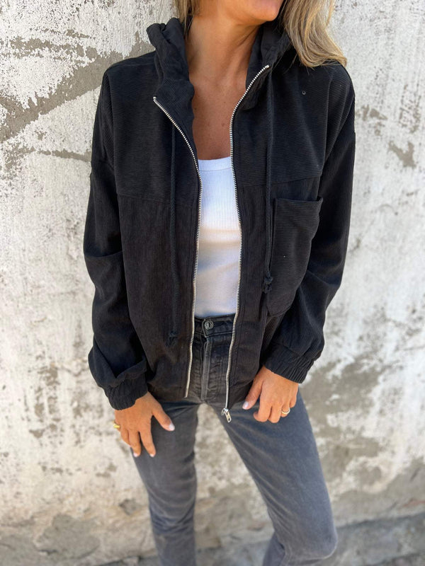 Vrouw in zwarte corduroy jas met capuchon, witte top en grijze jeans tegen betonnen muur. Modieuze herfstkleding, casual damesoutfit.