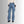 Vrouw in blauwe jeans met unieke golvende naden, staand tegen een grijze achtergrond. Modieuze damesbroek, casual stijl, trendy denim.
